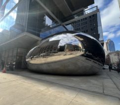 A large public sculpture on a New York City sidewalk consisting of polished stainless steel panels welded together to create a rounded, smooth surface reflecting and distorting the surrounding area.