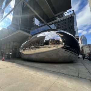 A large public sculpture on a New York City sidewalk consisting of polished stainless steel panels welded together to create a rounded, smooth surface reflecting and distorting the surrounding area.