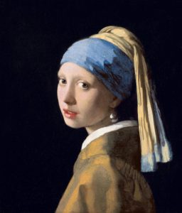 A portrait of a young woman looking towards the viewer over her left shoulder against a black background wearing a blue and yellow turban as well as a pearl earring.