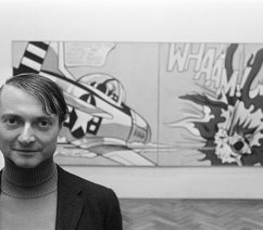 A black-and-white photograph of the pop artist Roy Lichtenstein standing in front of his work Whaam!, a comic book-style painting of a fighter plane.