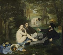 A group of figures in a wooded scene near water: two young men are fully dressed reclining on the grass, while the woman next to them is completely nude. Another women in the background is partially dressed while wading in the water behind them. A spread of food including breads and fruit is laid out beside the women's discarded clothes.