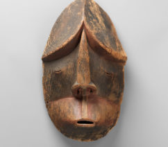 A carved wooden mask with small slits for the mouth and eyes created by the Alutiiq people of Alaska.