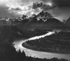 A black-and-white photograph of the winding Snake River in the foreground, with the Grand Teton mountains rising into the clouds in the background.