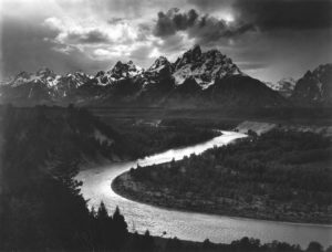 A black-and-white photograph of the winding Snake River in the foreground, with the Grand Teton mountains rising into the clouds in the background.