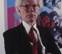 A color photograph of Andy Warhol in a suit and red tie standing in front of a colorful work featuring the faces of the Marx Brothers.