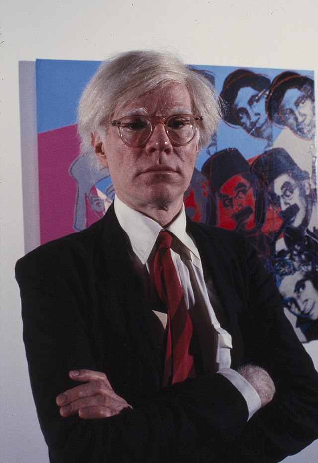 A color photograph of Andy Warhol in a suit and red tie standing in front of a colorful work featuring the faces of the Marx Brothers.