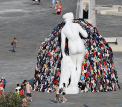 A large, public square featuring a white marble statue of a nude woman in front of a mound of dirty, multicolored clothing.