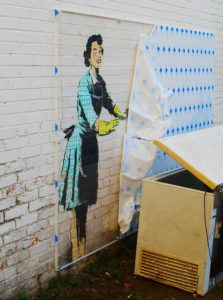 Graffiti of a woman in a green gingham dress and yellow rubber cloves on a white brick wall. The rest of the graffiti is covered by a tarp.