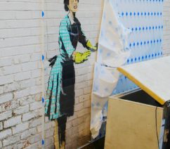 Graffiti of a woman in a green gingham dress and yellow rubber cloves on a white brick wall. The rest of the graffiti is covered by a tarp.
