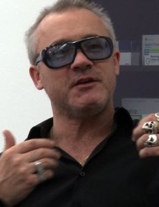 A photograph of the British artist Damien Hirst in a black polo shirt and sunglasses.