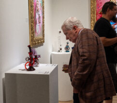 An older gentleman in a sportscoat and a drink inspecting a piece of functional glass sculpture on a pedestal.