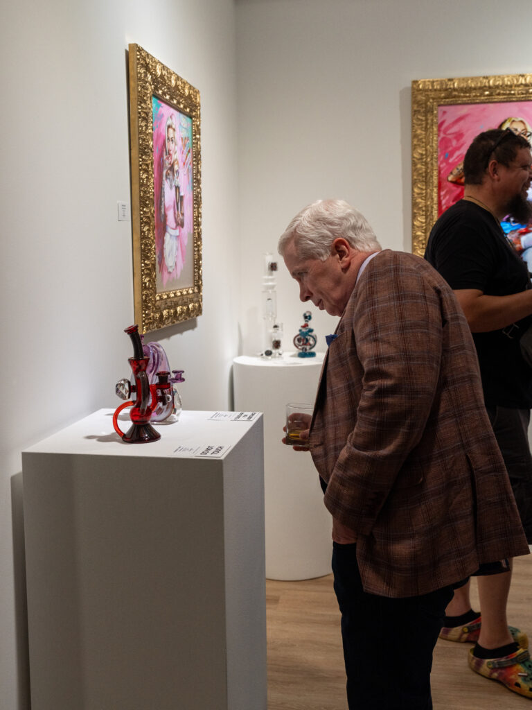 An older gentleman in a sportscoat and a drink inspecting a piece of functional glass sculpture on a pedestal.