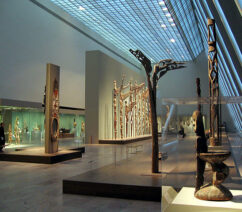 A large museum gallery of African and Oceanic artifacts lit through a glass ceiling.