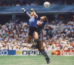 A photograph of Diego Maradona leaping into the air to punch a soccer ball out of the way of a goalkeeper.