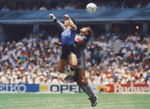 A photograph of Diego Maradona leaping into the air to punch a soccer ball out of the way of a goalkeeper.