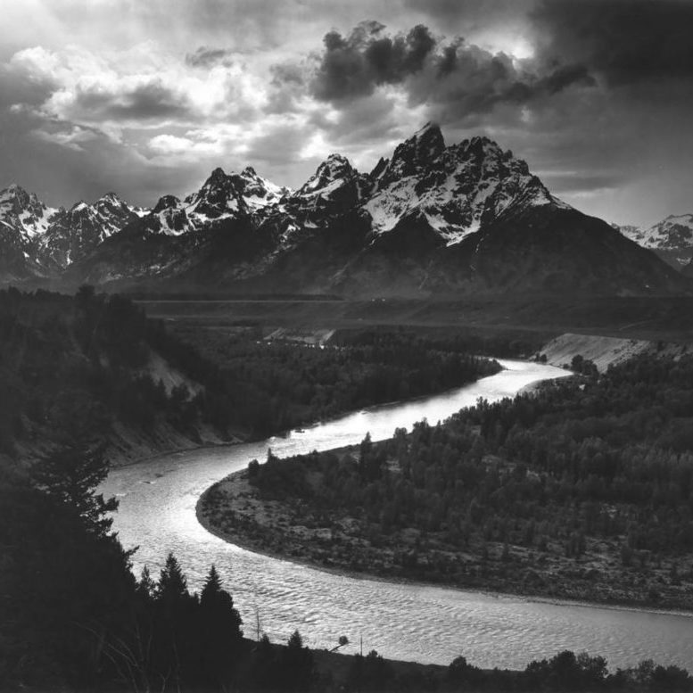 A black-and-white photograph of the winding Snake River in the foreground, with the Grand Teton mountains rising into the clouds in the background.