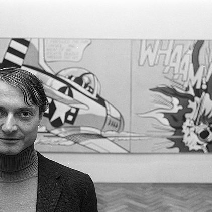 A black-and-white photograph of the pop artist Roy Lichtenstein standing in front of his work Whaam!, a comic book-style painting of a fighter plane.