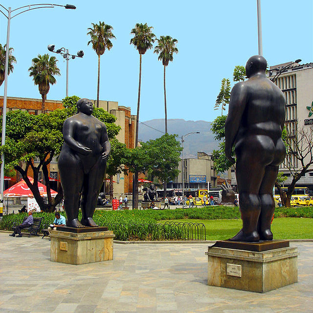Two bronze sculptures of a large nude man and a large nude woman woman facing one another in a public square.