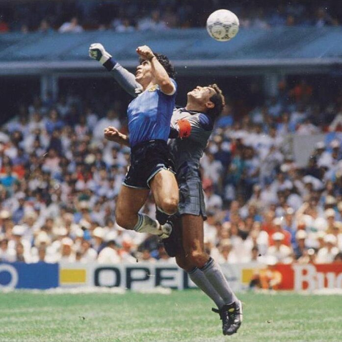 A photograph of Diego Maradona leaping into the air to punch a soccer ball out of the way of a goalkeeper.