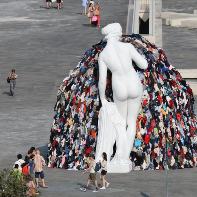A large, public square featuring a white marble statue of a nude woman in front of a mound of dirty, multicolored clothing.