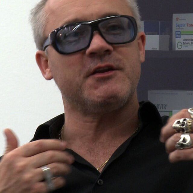 A photograph of the British artist Damien Hirst in a black polo shirt and sunglasses.