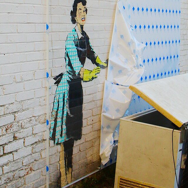 Graffiti of a woman in a green gingham dress and yellow rubber cloves on a white brick wall. The rest of the graffiti is covered by a tarp.