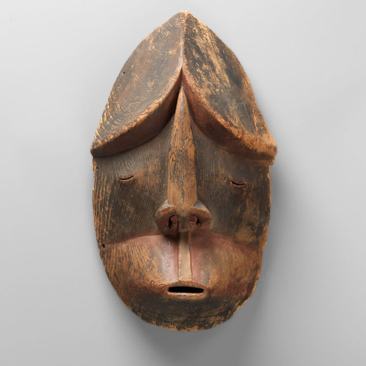 A carved wooden mask with small slits for the mouth and eyes created by the Alutiiq people of Alaska.